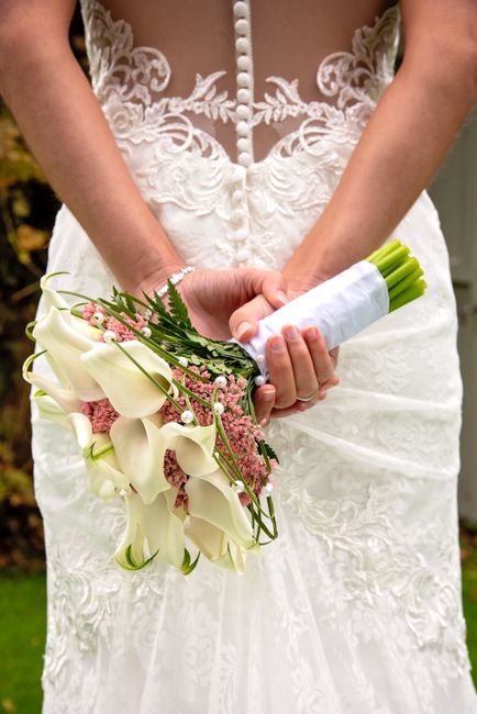 A close-up image of a bride’s hands holding a wedding bouquet, featuring white calla lilies, soft pink waxflowers, and green foliage, wrapped in white ribbon. The bride wears a white lace wedding go