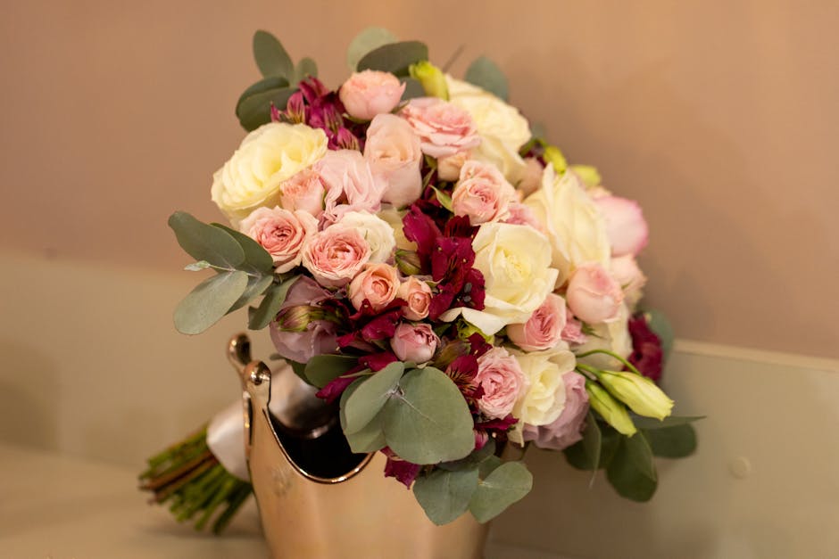 A bouquet arrangement featuring various fresh flowers including pale pink roses, creamy white roses, and deep burgundy blooms, accented with silvery-green eucalyptus leaves. The bouquet is crafted wit