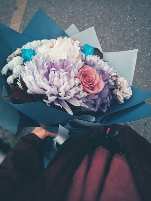 A hand holding a floral bouquet wrapped in blue paper, featuring a variety of fresh flowers including large white chrysanthemums, a soft pink rose, purple lisianthus, and small turquoise accents. The 